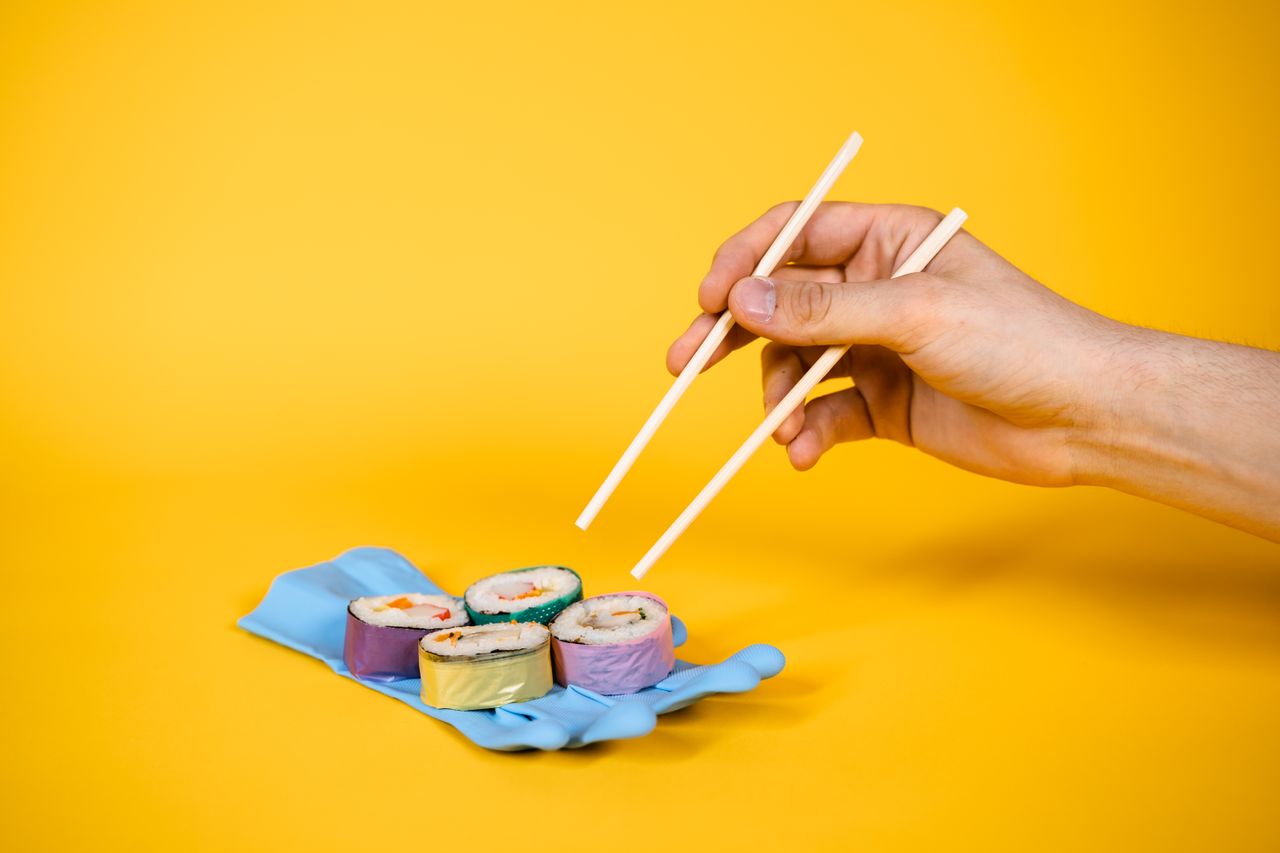Home Artistic sushi on a rubber glove with chopsticks, set against a vibrant yellow background.
